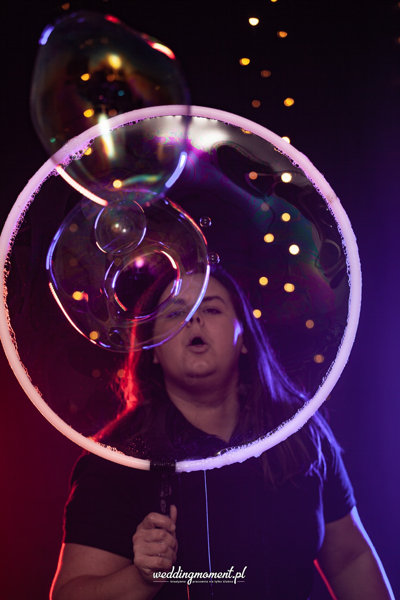 Kobieta dmucha duże bańki mydlane za pomocą okrągłego przyrządu na tle ciemnego pomieszczenia z punktowymi światłami, oświetlona czerwonym i niebieskim światłem.