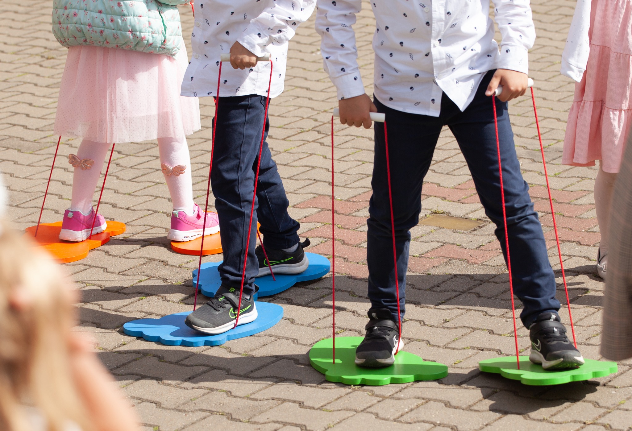 Grupa dzieci uczestniczy w zabawie na świeżym powietrzu, chodząc na kolorowych, drewnianych platformach przymocowanych do rąk za pomocą czerwonych sznurków.
