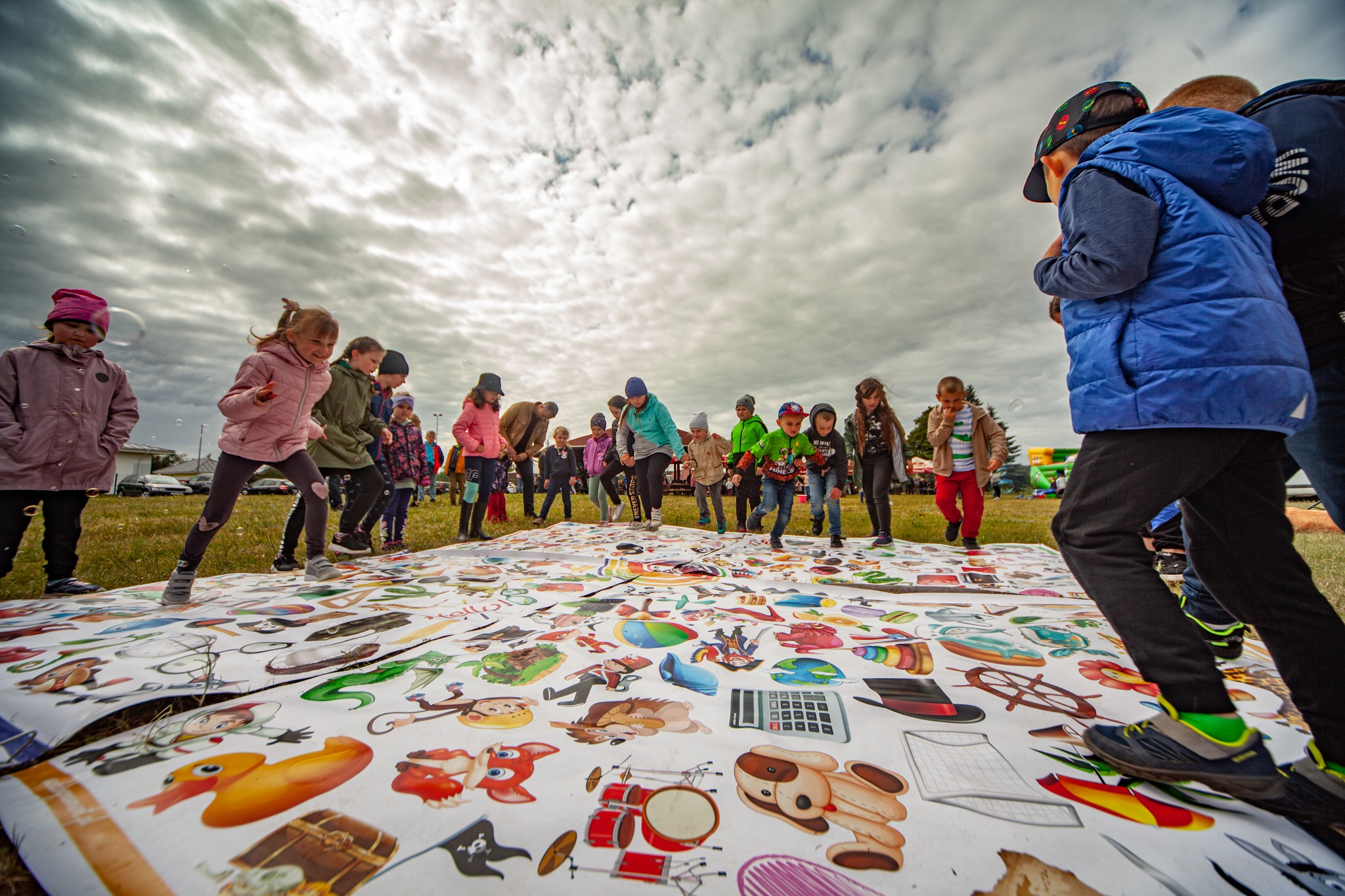 Grupa dzieci skacze po rozłożonej na trawie, dużej macie z ilustracjami, w tle widoczne pochmurne niebo i bańki mydlane.