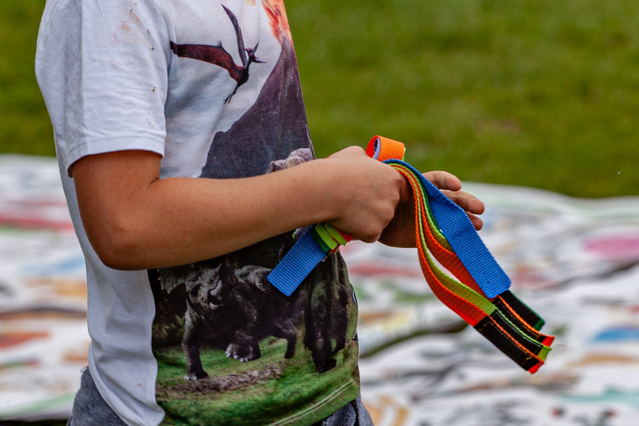 Chłopiec w koszulce z dinozaurami trzyma w dłoniach kolorowe, parciane paski z czarnymi zakończeniami na tle rozłożonej, malowanej płachty.