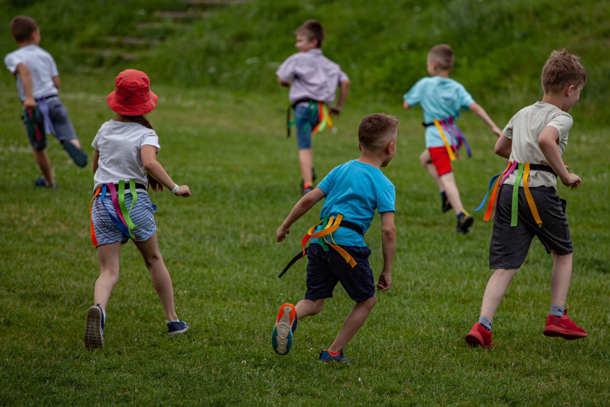 Grupa dzieci biegnie po trawie podczas zabawy w berka z kolorowymi wstążkami przyczepionymi do pasków.