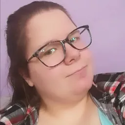 Portret kobiety w okularach z brązową oprawką, ubranej w koszulę w kratę, na tle fioletowej ściany.