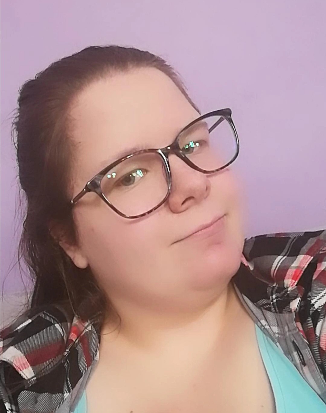 Portret kobiety w okularach z brązową oprawką, ubranej w koszulę w kratę, na tle fioletowej ściany.