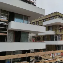 Budynek wielorodzinny w trakcie ocieplania elewacji, widoczne rusztowania, izolacja termiczna, częściowo zamontowane okna, oraz żuraw budowlany w tle.