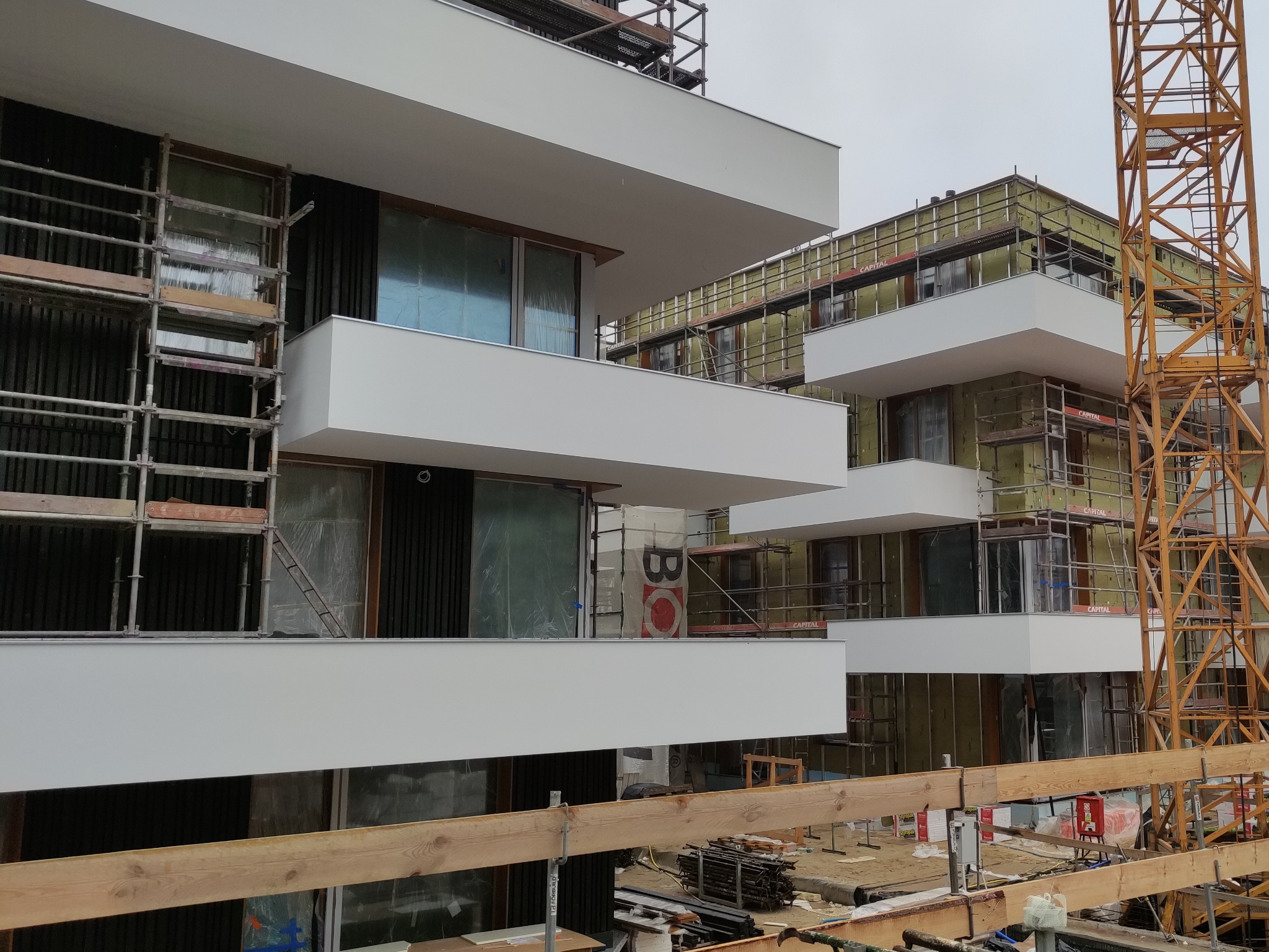 Budynek wielorodzinny w trakcie ocieplania elewacji, widoczne rusztowania, izolacja termiczna, częściowo zamontowane okna, oraz żuraw budowlany w tle.