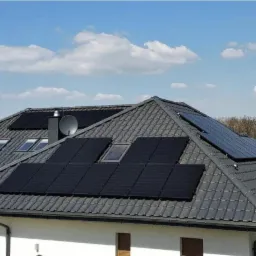 Czarny dach z szarej dachówki z panelami fotowoltaicznymi i oknami dachowymi, na tle błękitnego nieba z chmurami.