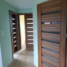 Wnętrze mieszkania z trzema zamontowanymi, drewnianymi drzwiami wewnętrznymi z poziomymi przeszkleniami, widoczne na tle zielonych ścian i podłogi z płytek imitujących drewno.