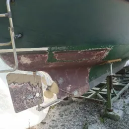 Spód łodzi przed piaskowaniem i malowaniem, widoczne ślady korozji i starej farby, łódź podparta na stalowej konstrukcji.