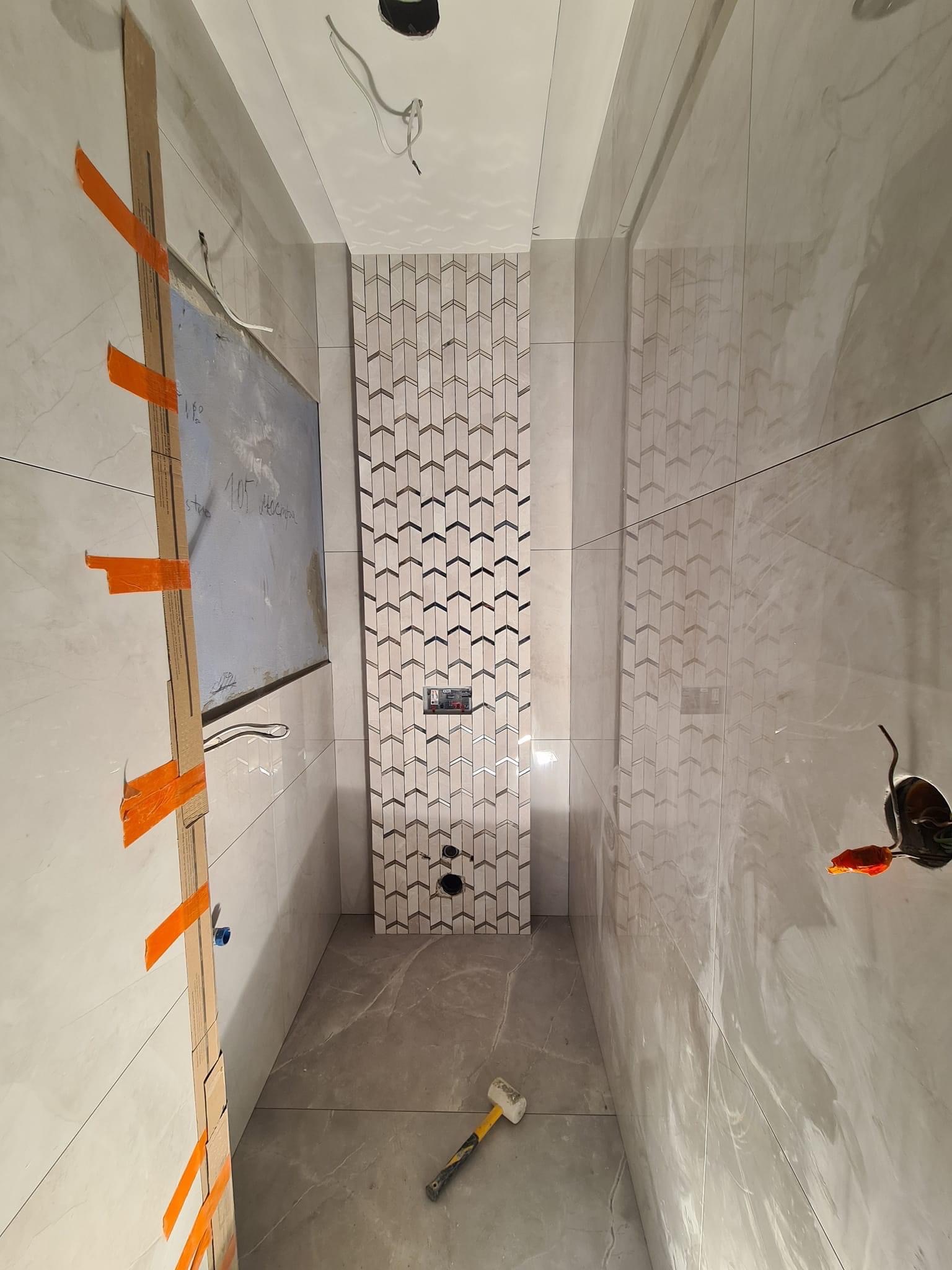 Wąska łazienka w trakcie remontu, z częściowo położonymi płytkami w geometryczny wzór na ścianie za toaletą, widoczne narzędzia glazurnicze i karton gips z notatkami.