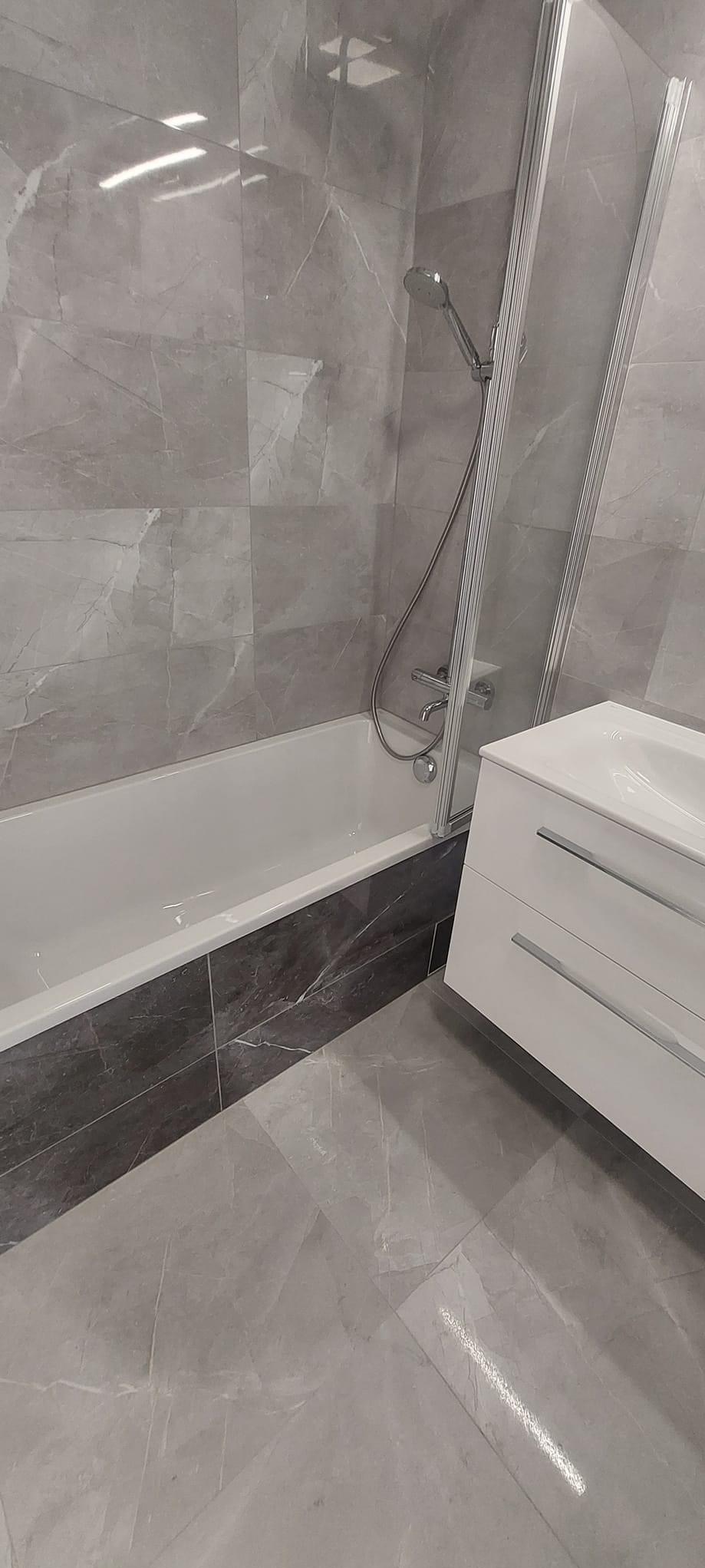 Wykończona łazienka z szarymi płytkami imitującymi marmur na ścianach i podłodze, biała wanna z ciemnoszarą obudową i szklanym parawanem, biała szafka pod umywalką z dwoma szufladami.