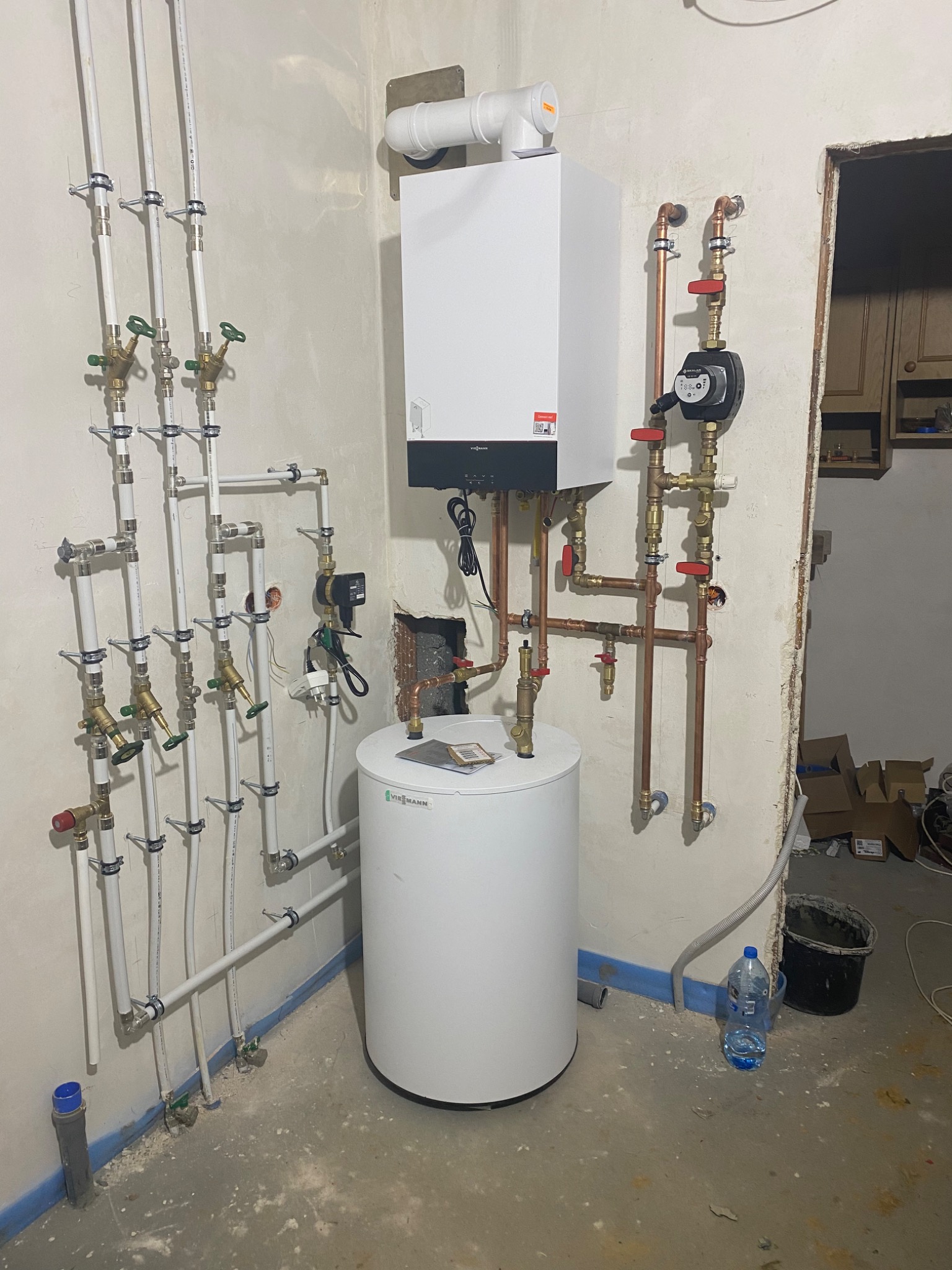 Biały piec grzewczy Viessmann z podłączonymi miedzianymi i plastikowymi rurami, widoczny zbiornik na wodę, instalacja w surowym pomieszczeniu z betonową podłogą i ścianami w trakcie wykończenia.