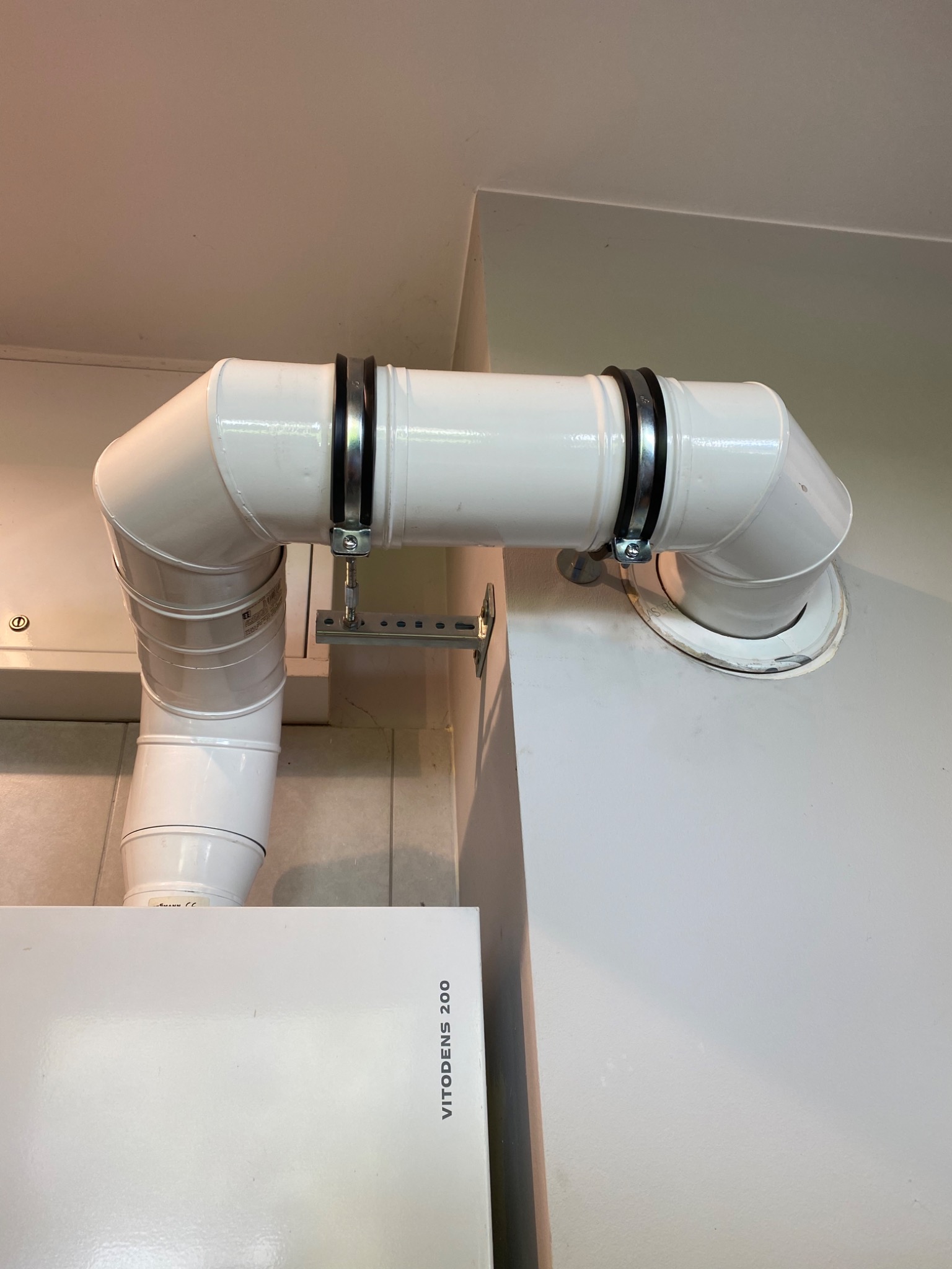 Białe rury wentylacyjne z metalowymi obejmami, podłączone do pieca Vitodens 200, widoczne elementy montażowe do ściany.