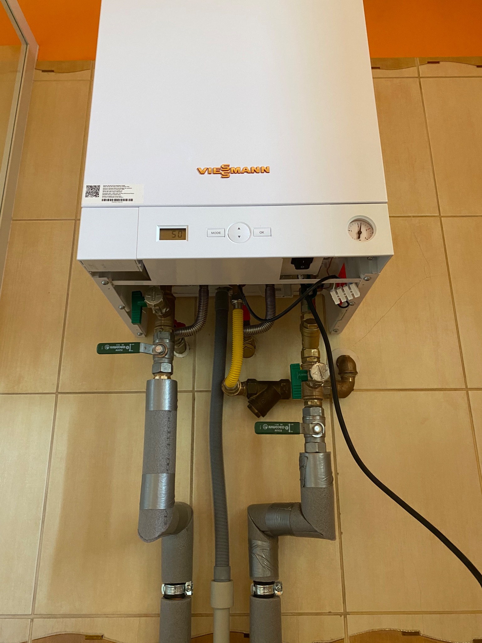 Biały piec grzewczy Viessmann zamontowany na ścianie z beżowych płytek, widoczne przyłącza hydrauliczne i gazowe, izolacja rur i przewody elektryczne.