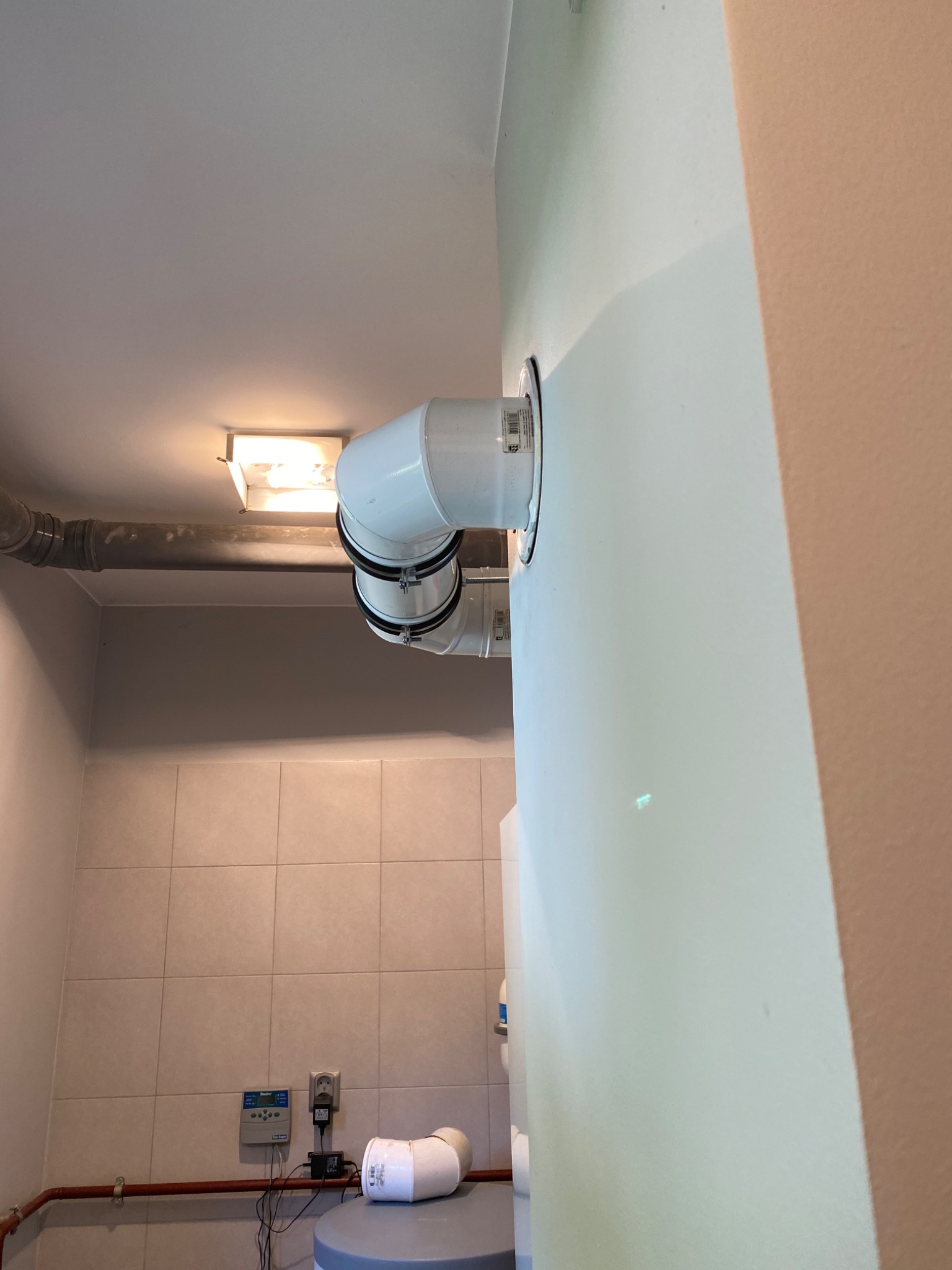 Białe rury wentylacyjne z czarnymi zaciskami przymocowane do ściany w pomieszczeniu z płytkami, widoczna lampa sufitowa i rura w tle.