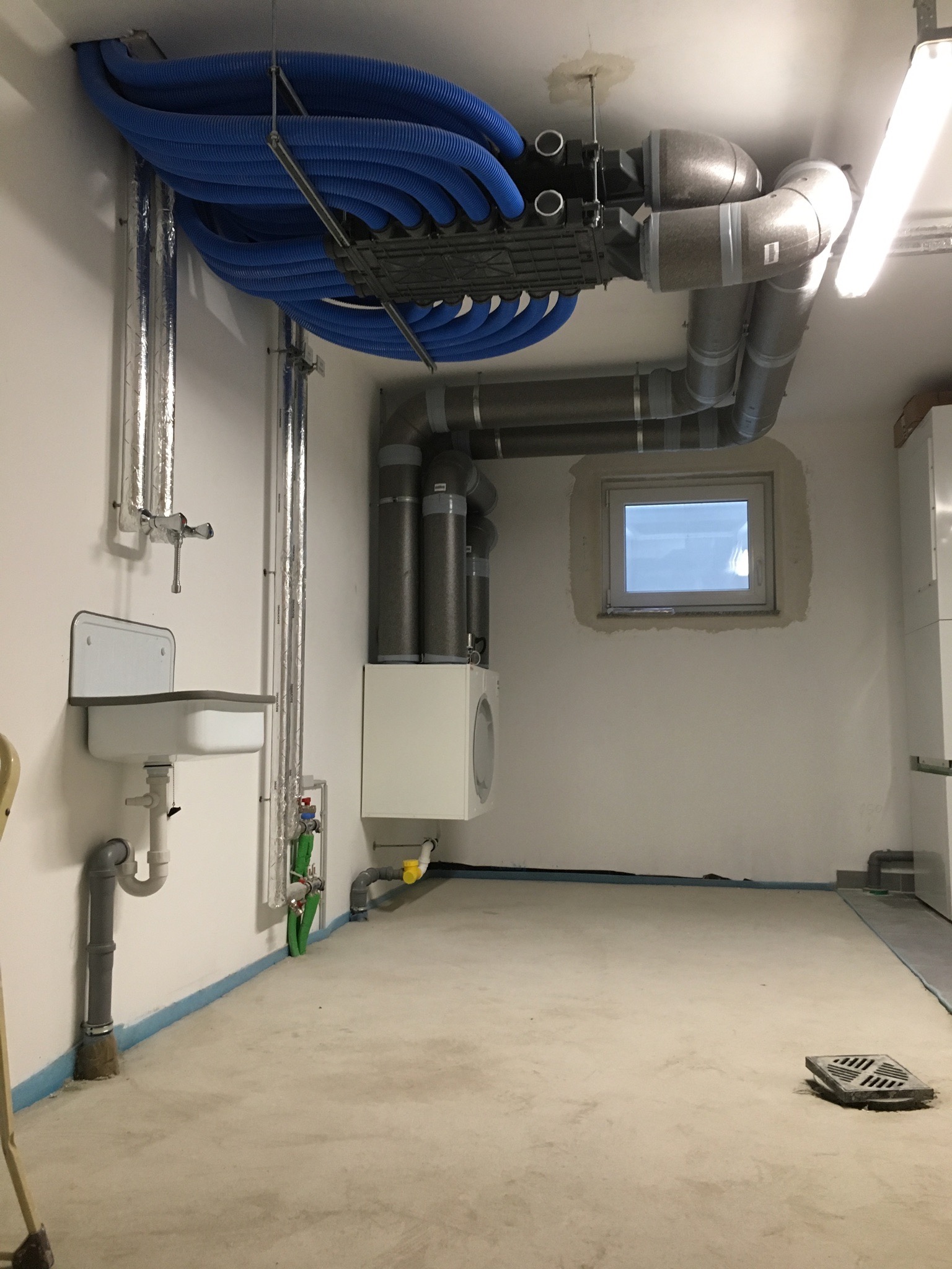 Instalacja rekuperacji z niebieskimi przewodami wentylacyjnymi i centralą pod sufitem w pomieszczeniu technicznym w Zalas, widoczny także zlew i okno.