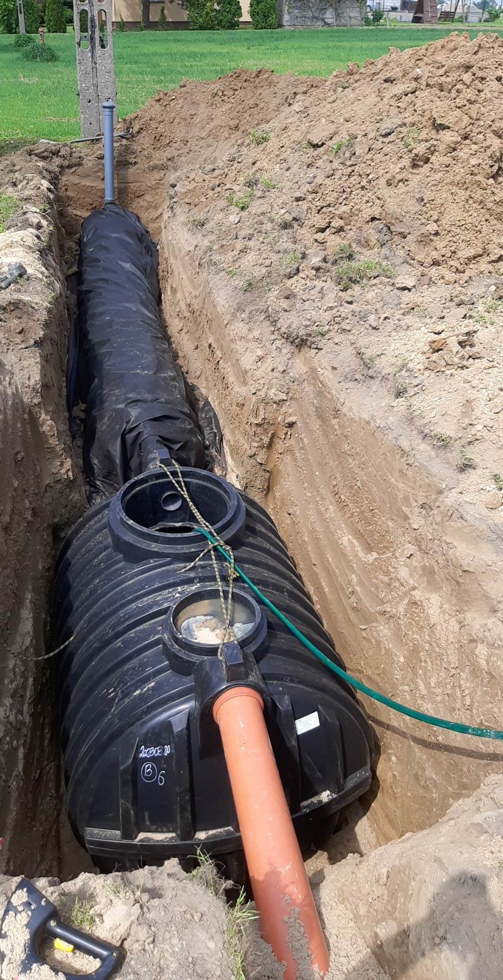 Czarny zbiornik na szambo umieszczony w wykopie z widoczną rurą odpływową, zielonym wężem i kablem oraz narzędziem do kopania obok.