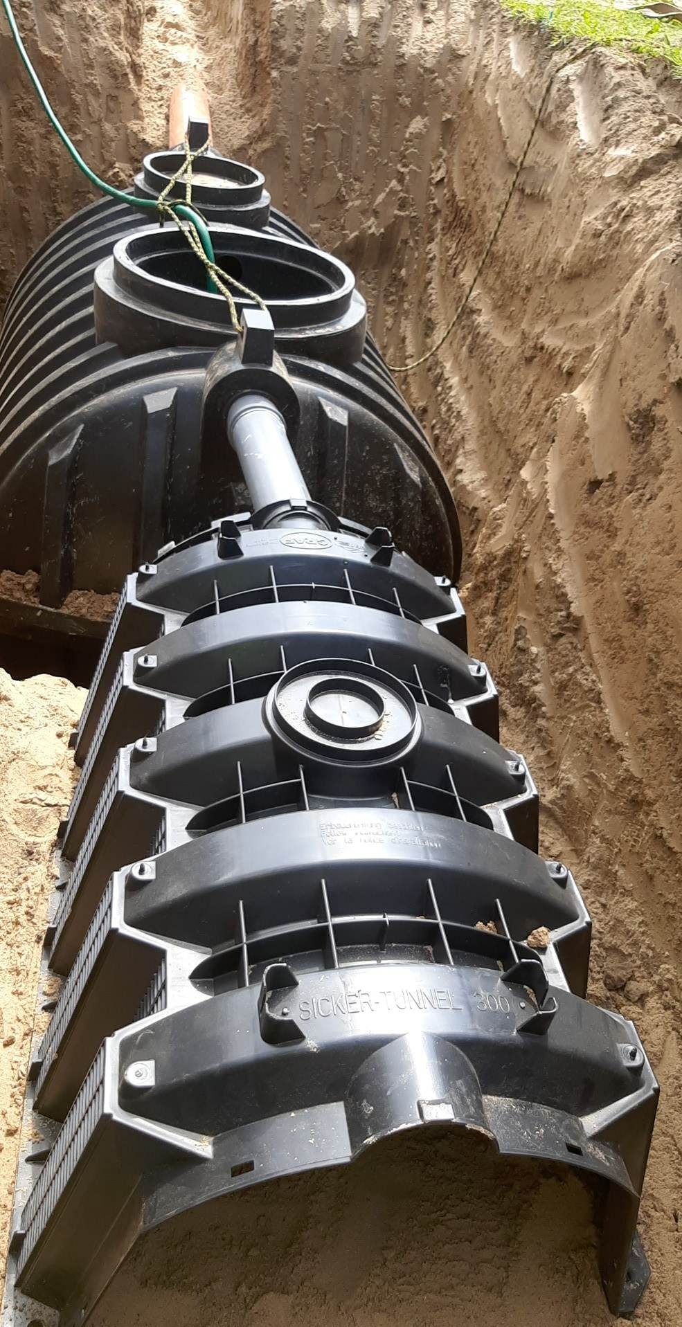 Czarny, modułowy tunel rozsączający Sicker-Tunnel 300, częściowo zakopany w wykopie z piasku, połączony z rurą kanalizacyjną, widok z góry.