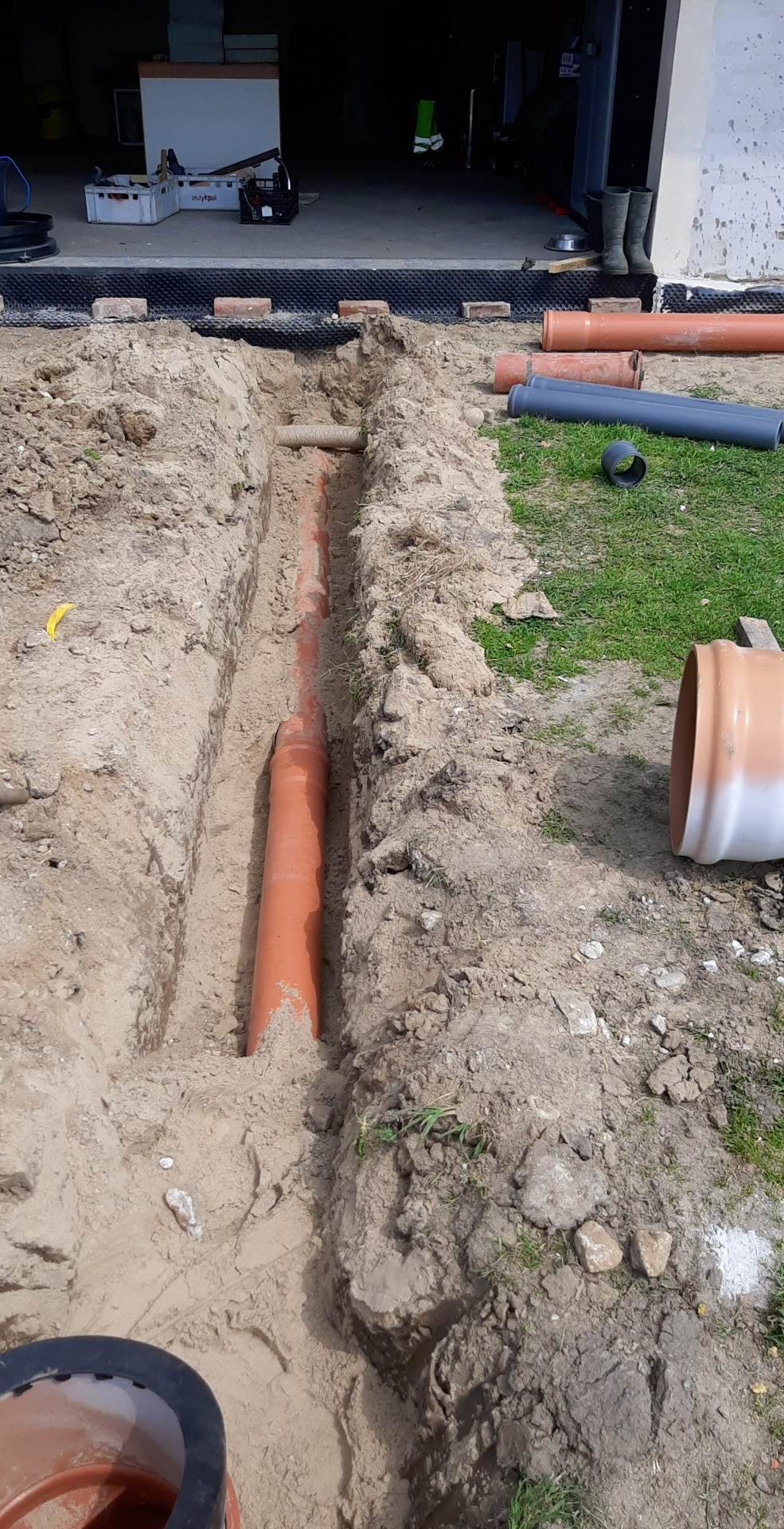 Wąski wykop w piaszczystej ziemi z ułożoną pomarańczową rurą kanalizacyjną, widoczne luźno leżące rury i fragment trawnika. W tle budynek gospodarczy z gumowcami przy wejściu.