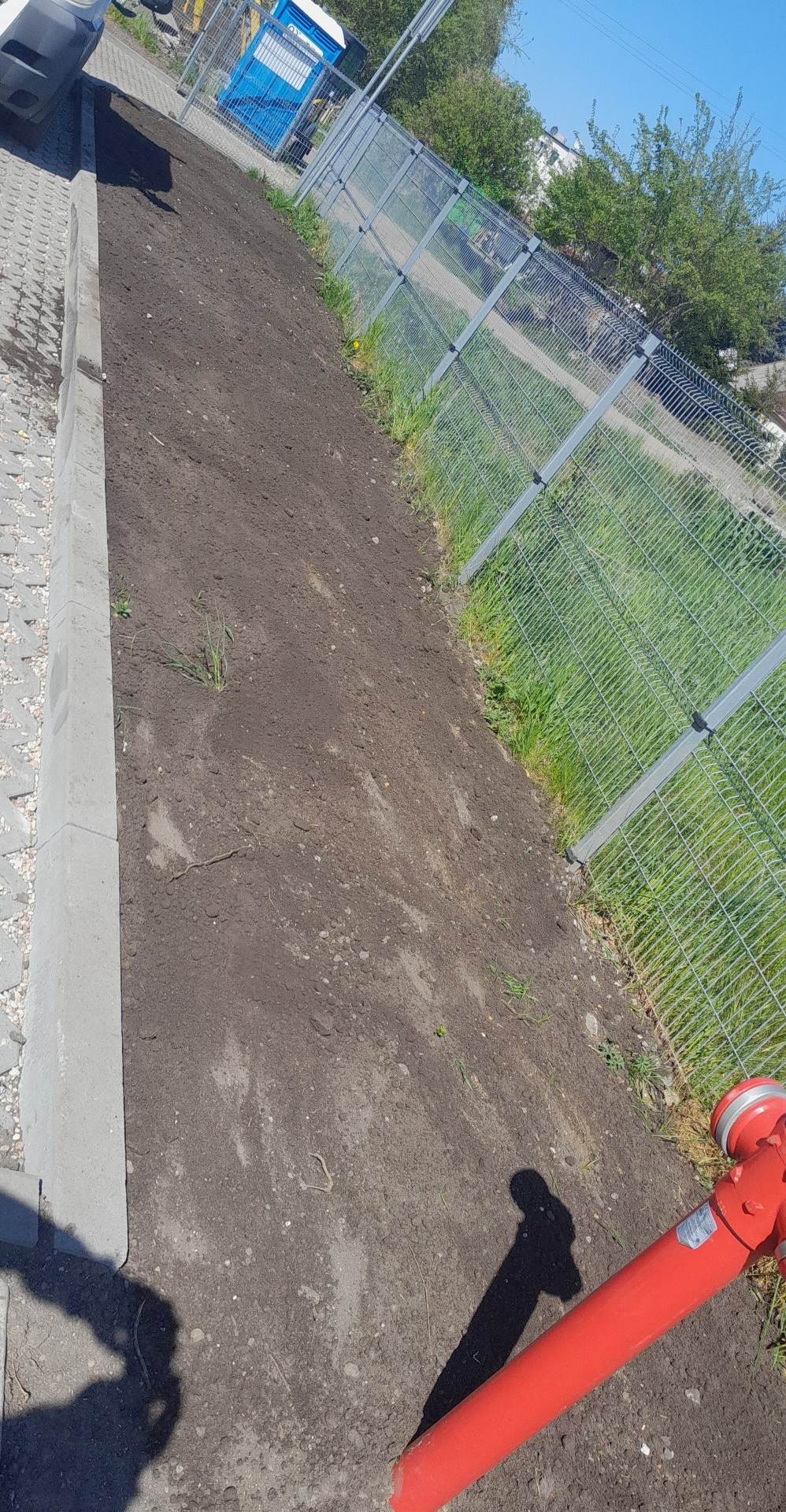 Przygotowany teren pod trawnik lub nasadzenia, z widocznym hydrantem przeciwpożarowym i ogrodzeniem z siatki.
