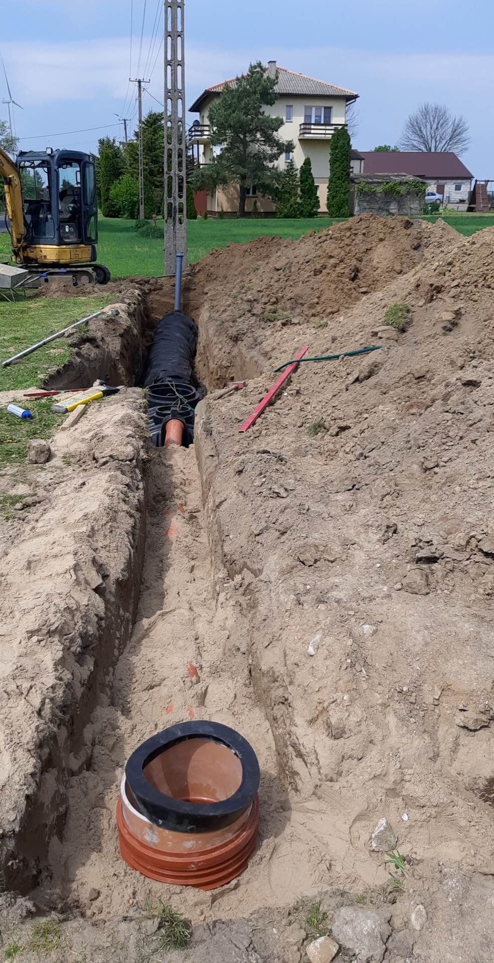 Wykopany rów w ziemi z ułożonymi rurami kanalizacyjnymi i studzienką inspekcyjną na pierwszym planie, w tle widoczny mały żółty koparka i dom.