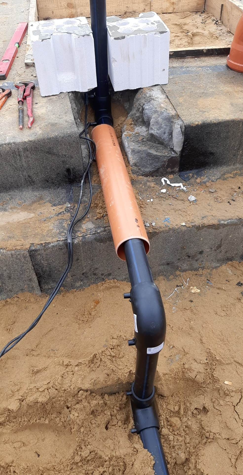 Czarna rura instalacyjna, częściowo osłonięta pomarańczową rurą, prowadząca przez wykop w ziemi, zabezpieczona bloczkami betonowymi, widoczne narzędzia budowlane w tle.