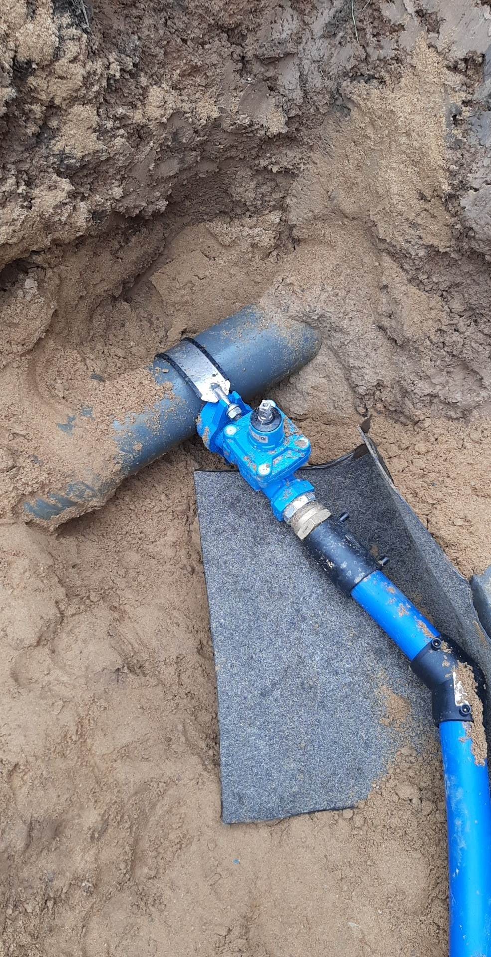 Szary rurociąg z niebieskim zaworem i niebieską rurą w wykopie z piaskiem, widoczna szara mata ochronna.