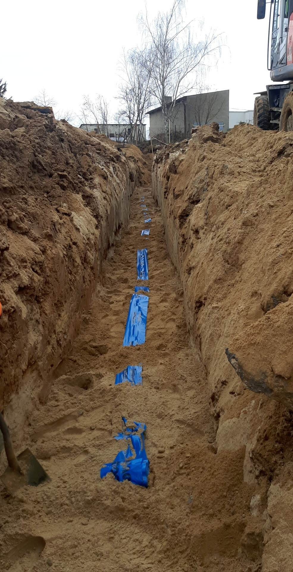 Wąski wykop w piaszczystej ziemi, zabezpieczony niebieską folią w regularnych odstępach, z widocznym fragmentem koparki na górze i narzędziem w lewym dolnym rogu.