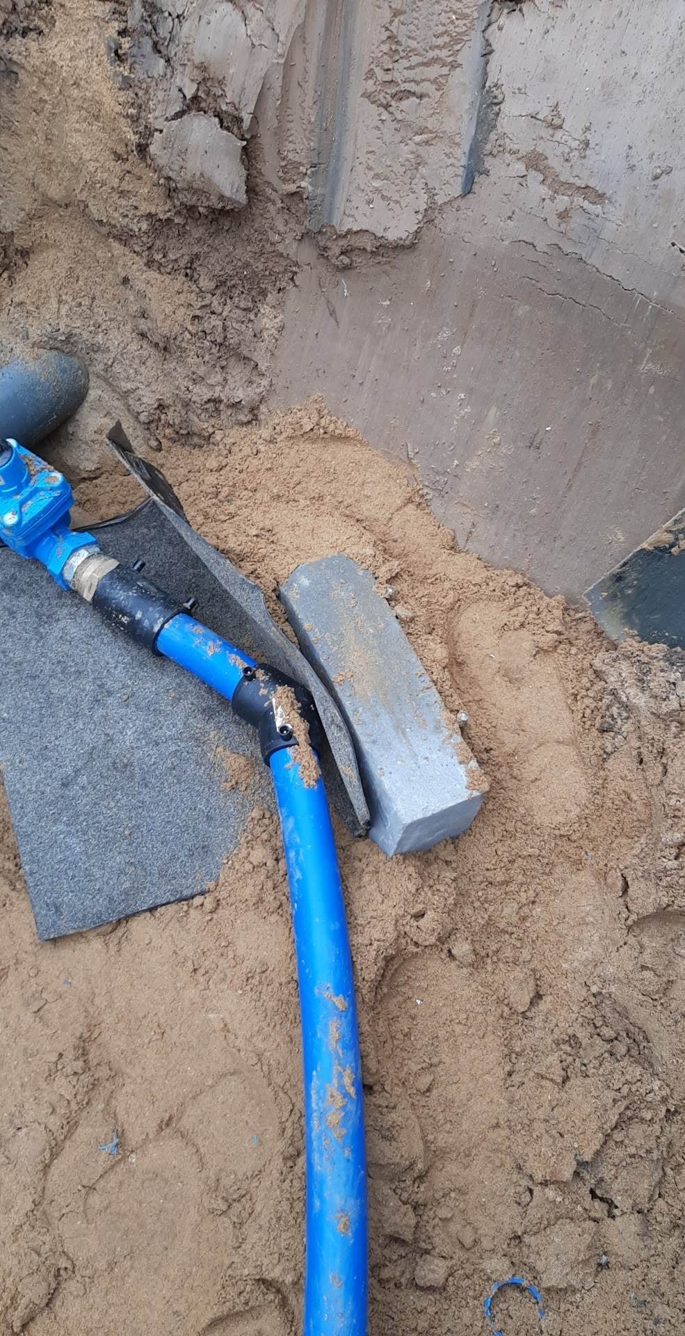 Niebieska rura instalacyjna z łącznikami, ułożona w wykopie wypełnionym piaskiem, obok fragmentu kostki brukowej i geowłókniny.