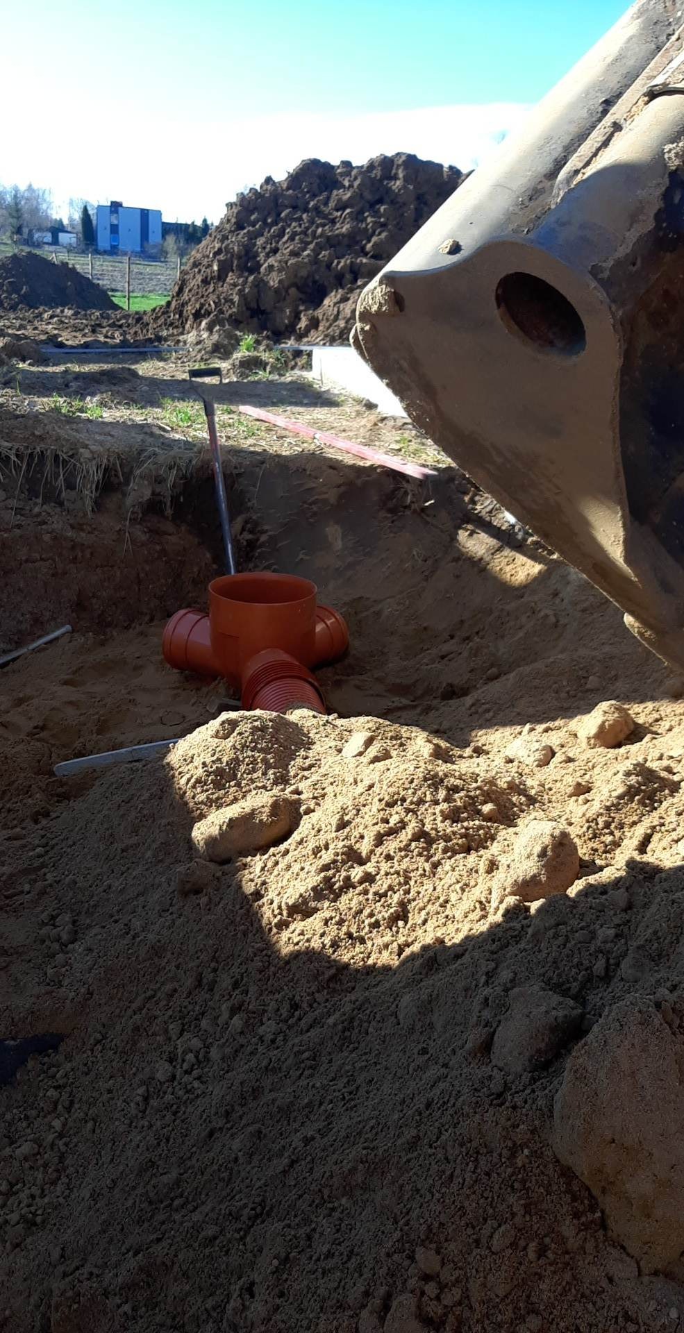 Pomarańczowe rury kanalizacyjne ułożone w wykopie obok łyżki koparki, w tle pryzma ziemi i budynek.