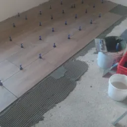 Układanie płytek podłogowych imitujących drewno z użyciem niebieskich klipsów poziomujących; widoczny klej z grzebieniem, wiadro z zaprawą i narzędzia.