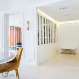 Elegancka, biała zabudowa z przeszklonymi szafkami i złotymi uchwytami, uzupełniona złotą ławką z białym siedziskiem, widoczna w jasnym wnętrzu z parkietem ułożonym w jodełkę i złotym krzesłem...