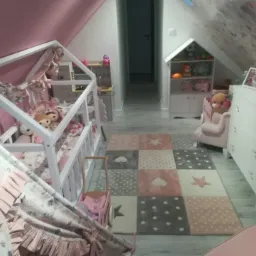 Pokój dziecięcy na poddaszu z łóżkiem w kształcie domku, tipi z falbanami i dywanem w geometryczne wzory, utrzymany w odcieniach bieli i różu.