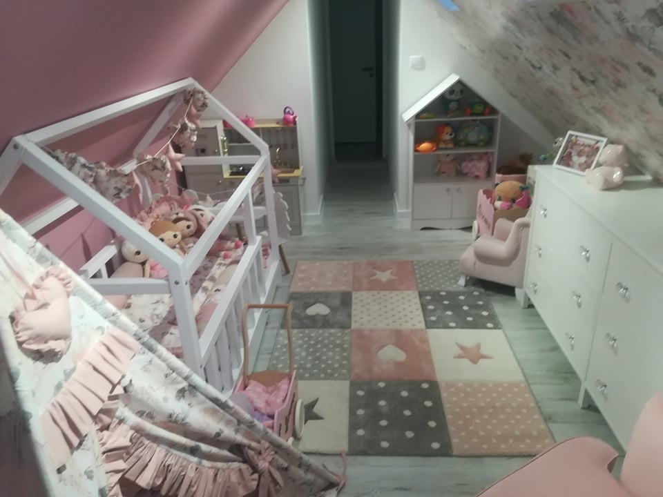 Pokój dziecięcy na poddaszu z łóżkiem w kształcie domku, tipi z falbanami i dywanem w geometryczne wzory, utrzymany w odcieniach bieli i różu.