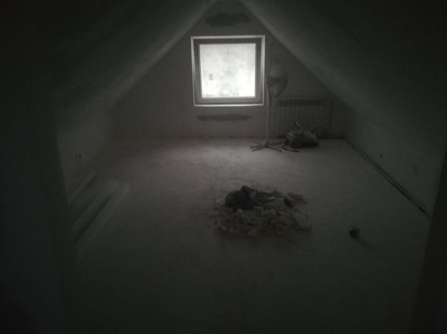 Poddasze ze skośnym sufitem w trakcie remontu, widoczne okno, grzejnik, wentylator stojący i stos gruzu na podłodze.