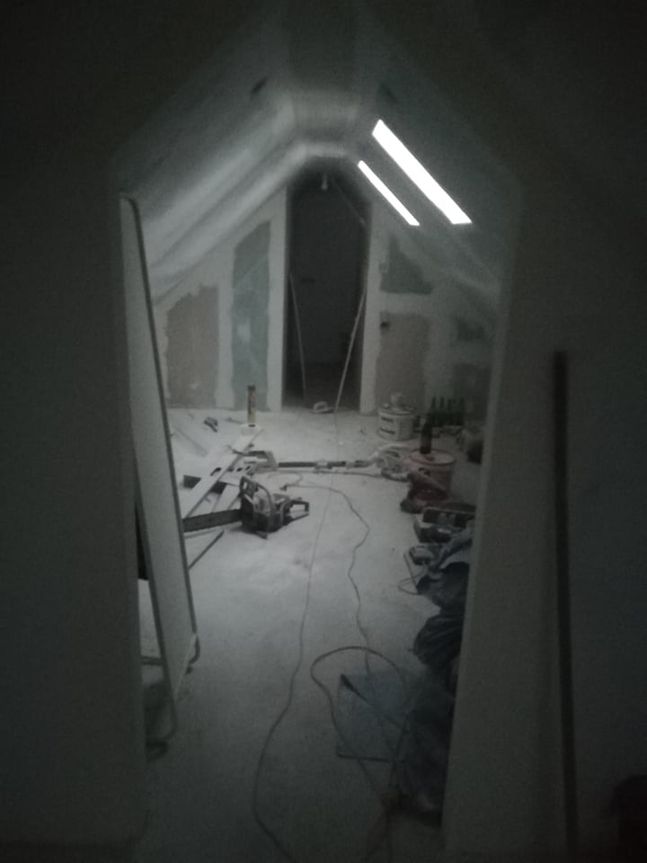 Poddasze w trakcie prac wykończeniowych: widoczne płyty gipsowo-kartonowe na skosach, narzędzia i materiały budowlane na podłodze, przez otwarte drzwi widoczne kolejne pomieszczenie, słabe...