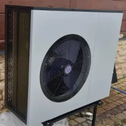 Zewnętrzna jednostka urządzenia grzewczego z wentylatorem, zamontowana na metalowym stelażu, na tle ściany z kamienia i drewnianej elewacji.