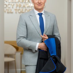 Kancelaria Radcy Prawnego Paweł Tołoczko - Mężczyzna w jasnoszarym garniturze i niebieskim krawacie, trzymający czarną togę z niebieskimi akcentami, stoi w nowoczesnym wnętrzu kancelarii.