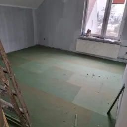 Przygotowanie podłoża pod ogrzewanie podłogowe w pokoju ze skośnym sufitem, widoczna drabina malarska i okno z grzejnikiem.