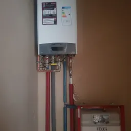 Zainstalowany, biały piec gazowy Ariston z widocznymi rurami miedzianymi i zaworami, podłączony do instalacji wodnej i grzewczej w pomieszczeniu o beżowych ścianach.