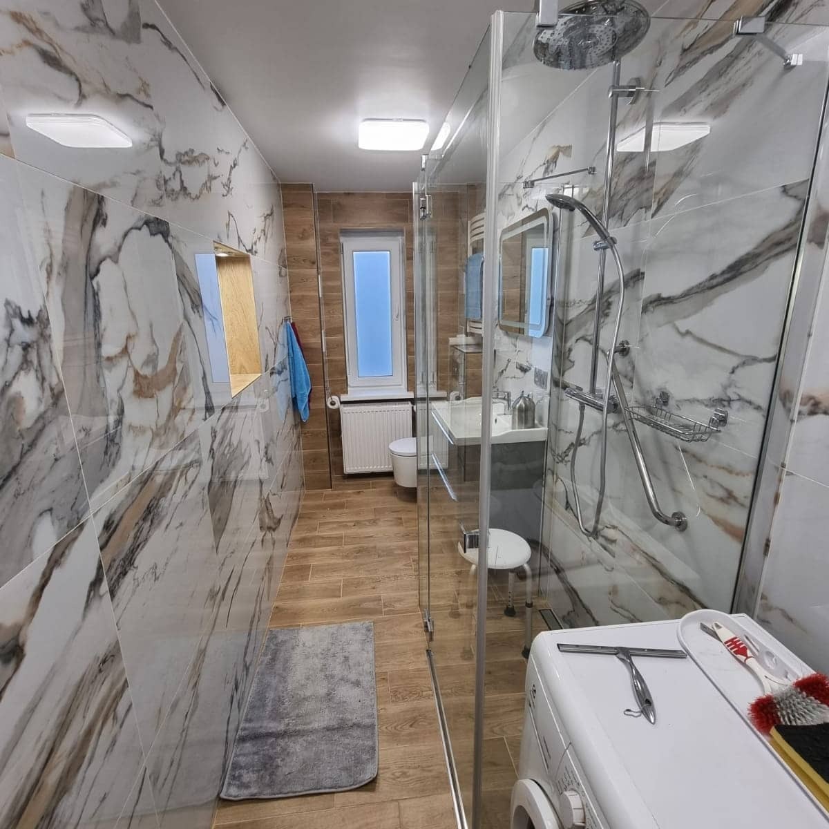 Wykończona łazienka w Wągrowcu z kabiną prysznicową typu walk-in, płytki imitujące marmur na ścianach, drewnopodobna podłoga oraz widoczne akcesoria łazienkowe.