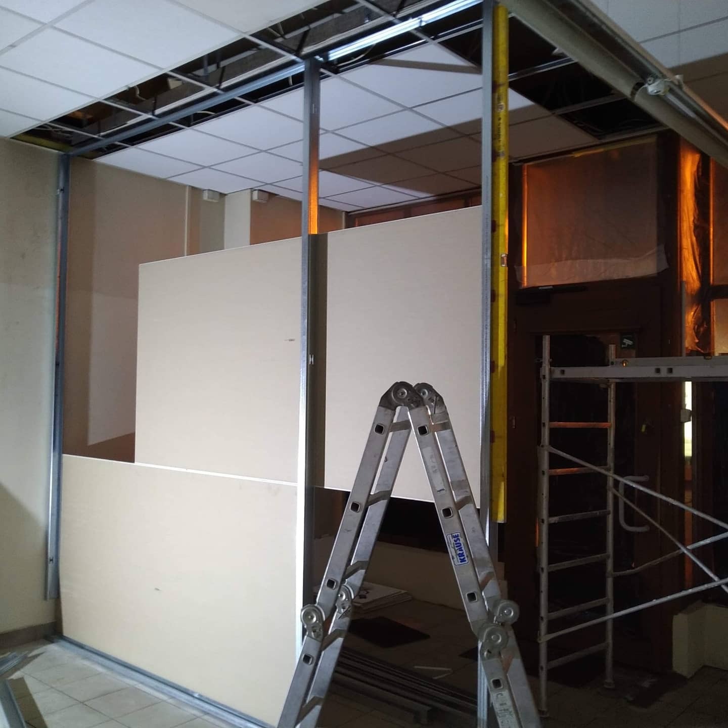 Montaż ścianki działowej z płyt gipsowo-kartonowych na metalowym stelażu, widoczna drabina Krause i rusztowanie w tle.
