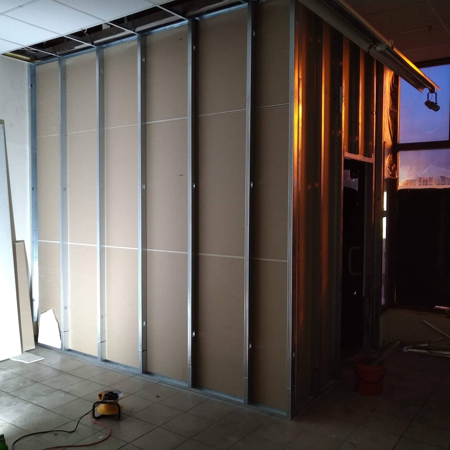 Stelaż z profili metalowych i płyt gipsowo-kartonowych w trakcie montażu, widoczne niedociągnięte płyty i prowizoryczne oświetlenie halogenowe.