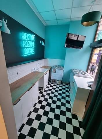 Wnętrze kuchni z czarno-białą podłogą w szachownicę, białymi szafkami z drewnianym blatem, neonowym napisem na ścianie i turkusowymi ścianami.