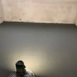 Świeżo wylana, szara posadzka jastrychowa w pomieszczeniu, widoczna maszyna do zacierania w dolnej części kadru, ściany przygotowane do malowania.