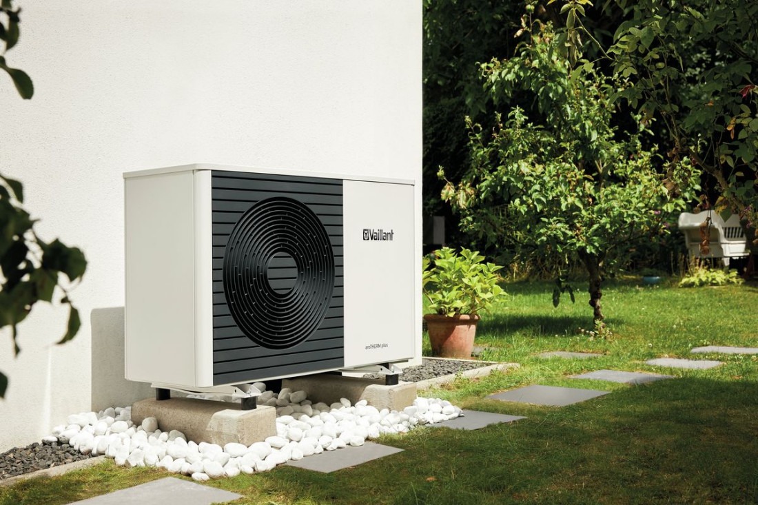 Biała pompa ciepła Vaillant aroTHERM plus, zamontowana na betonowych podstawach i otoczona białymi kamykami, na tle zielonego trawnika i ściany budynku.