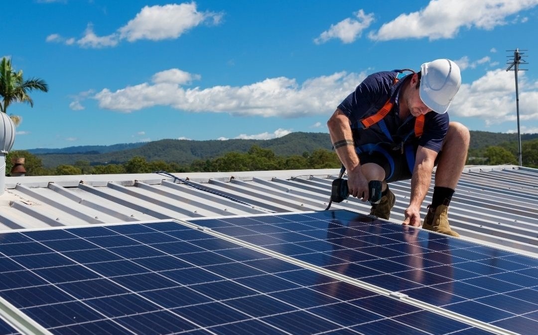 Monter w kasku i uprzęży instaluje panele słoneczne na blaszanym dachu, w tle zielone wzgórza i błękitne niebo z chmurami.