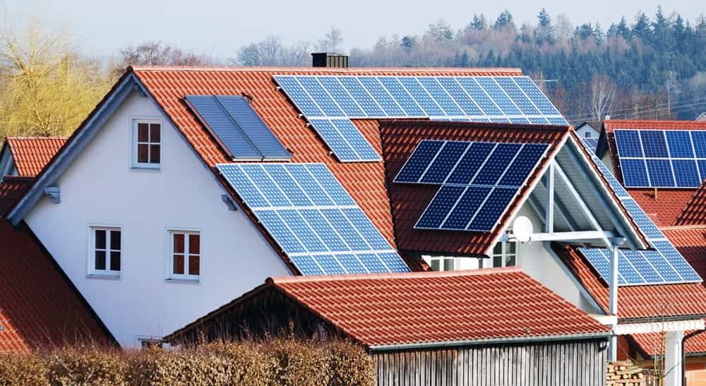 Biały dom jednorodzinny z czerwoną dachówką, na której zamontowane są panele słoneczne w różnych konfiguracjach, niektóre ustawione pionowo, inne poziomo. W tle widoczne drzewa.