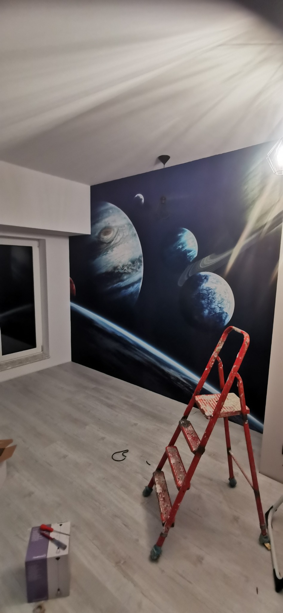 Pokój z fototapetą przedstawiającą planety i galaktyki, w tle widoczna drabina malarska i pudełko z narzędziami.