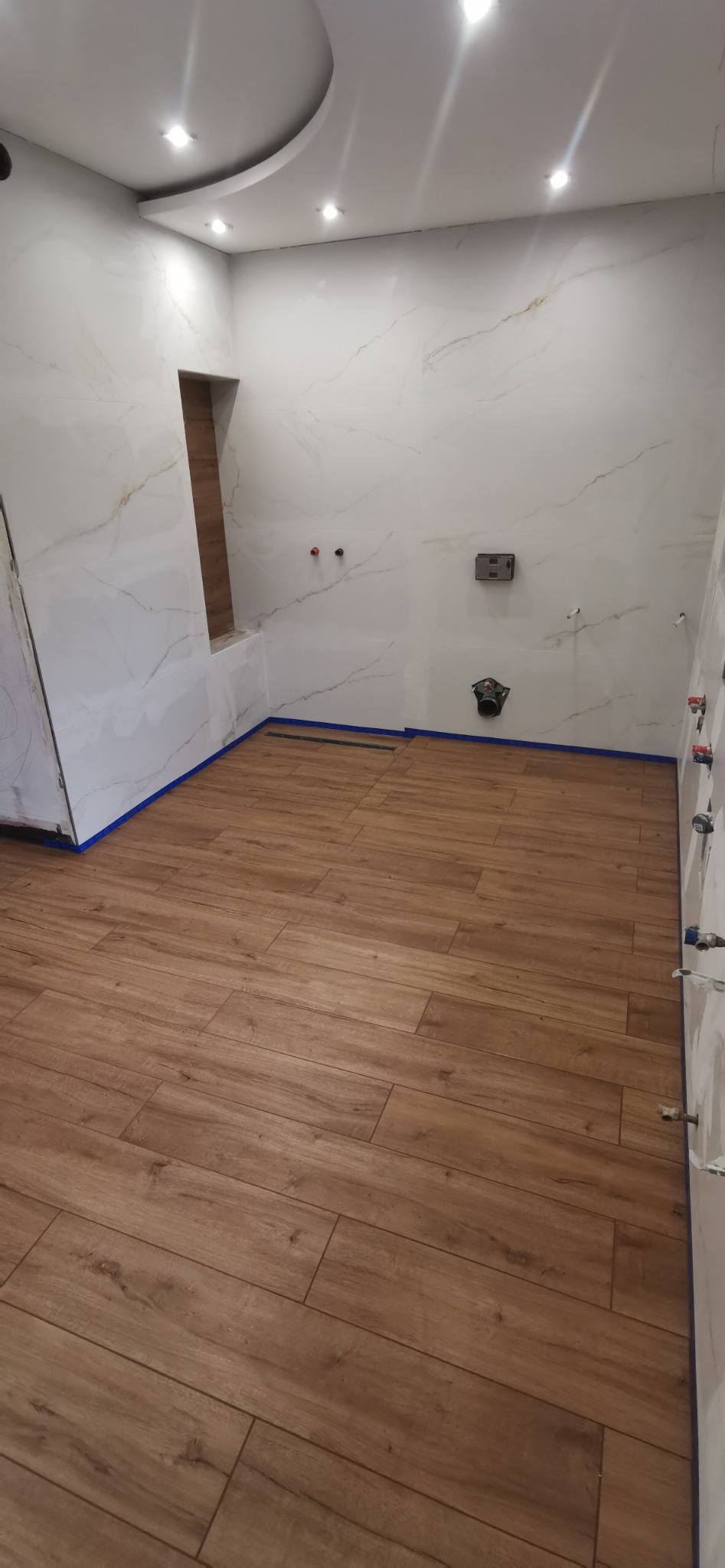 Łazienka w trakcie wykańczania: brązowe płytki imitujące drewno na podłodze, białe płytki marmurowe na ścianach z wnęką, zabezpieczone niebieską taśmą malarską, widoczne punkty instalacji wodnej.