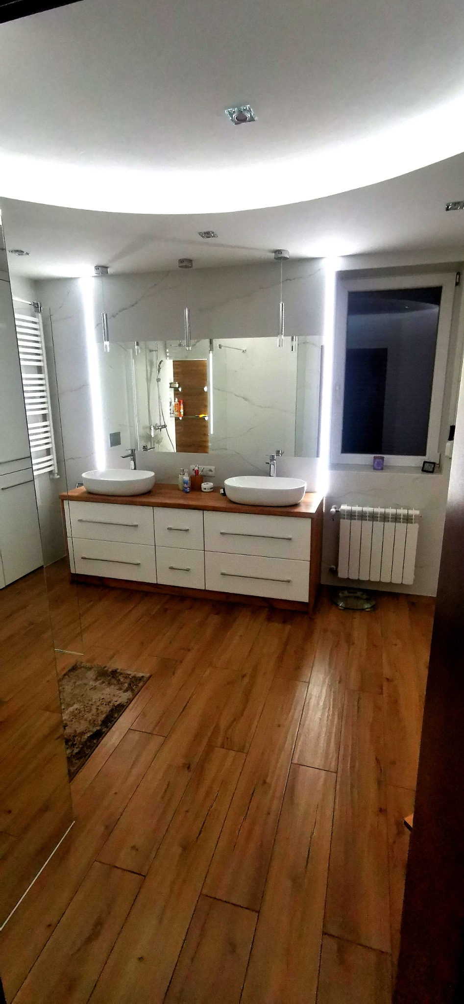 Nowoczesna łazienka z drewnianą podłogą, białymi szafkami pod umywalki i dużym lustrem z podświetleniem LED, widoczny grzejnik i dywanik.